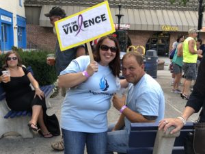 a man in an AquaPhoenix shirt and a woman in an AquaPhoenix shirt holding a sign to stop violence at the 2018 Walk A Mile In Her Shoes event